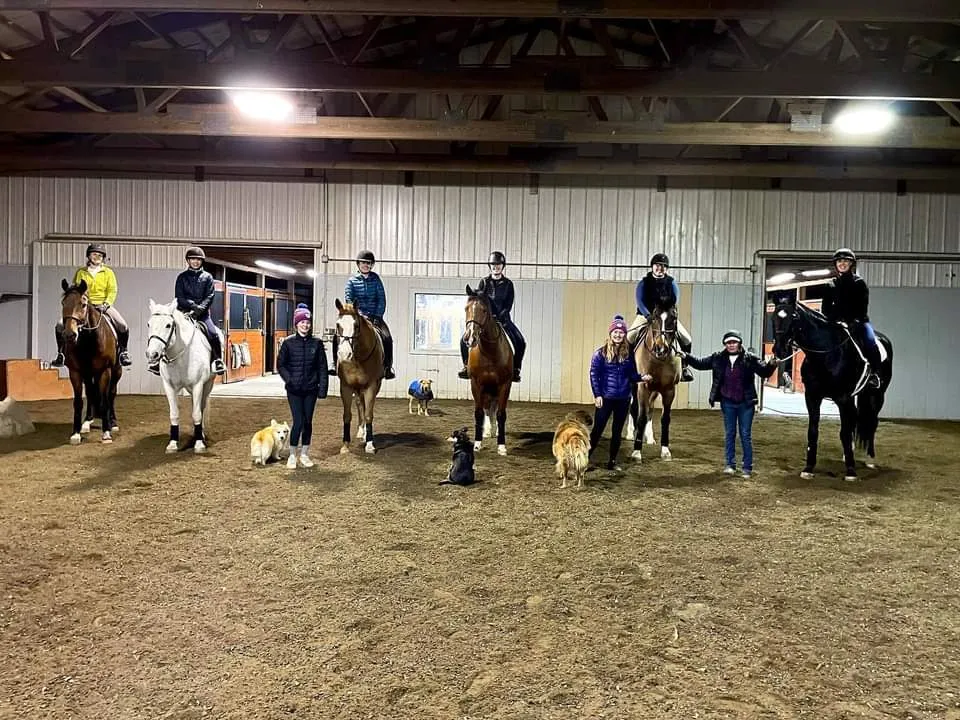 Group of riders on horseback with instructors and dogs in the indoor arena